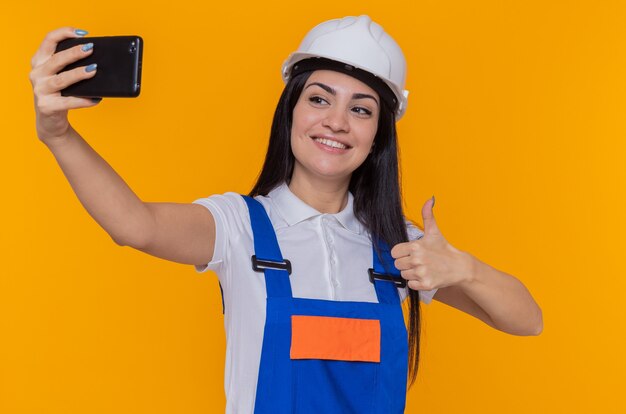 Young builder woman in construction uniform and safety helmet doing selfie using smartphone smiling confident showing thumb up standing over orange wall