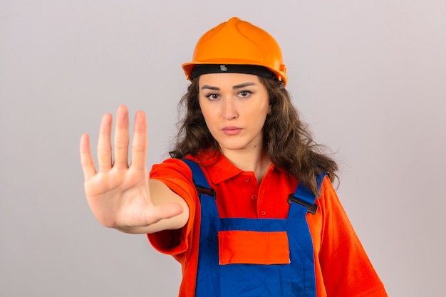 Молодая женщина строитель в строительной форме и защитный шлем делает стоп петь с ладонью выражения предупреждения руки над изолированной белой стеной
