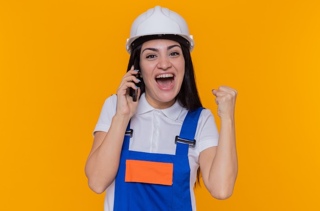 Young builder woman in construction uniform and safety helmet happy and excited clenching fist while talking on mobile phone standing over orange wall