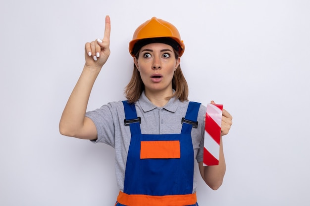 Free photo young builder woman in construction uniform and safety helmet holding adhesive tape looking aside surprised and worried showing index finger