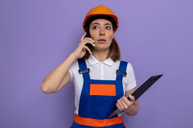 Young builder woman in construction uniform and safety helmet holding clipboard looking worried while talking on mobile phone standing over purple wall