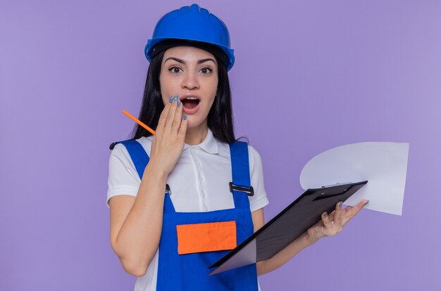 Young builder woman in construction uniform and safety helmet holding clipboard and pencil looking at front amazed and surprised standing over purple wall