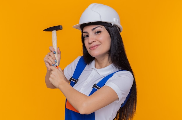 Young builder woman in construction uniform and safety helmet holding hammer looking at front with smile on face standing over orange wall