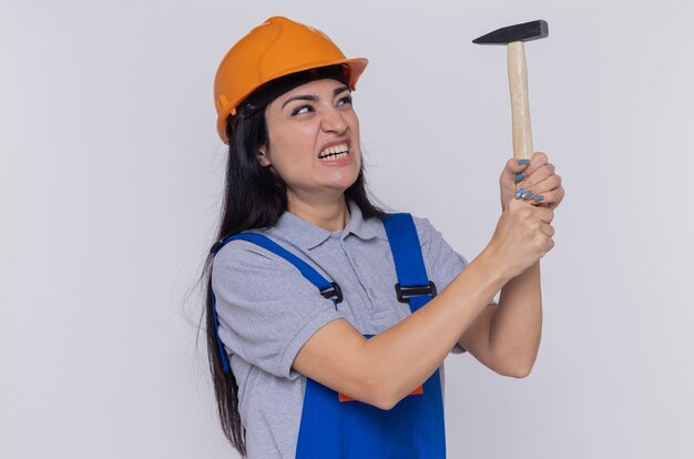 Young builder woman in construction uniform and safety helmet holding hammer yelling with annoyed expression standing over white wall