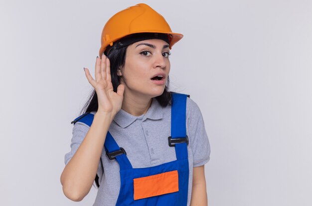 白い壁の上に立っているゴシップを聴こうとしている彼女の耳に手をつないで建設制服と安全ヘルメットの若いビルダーの女性