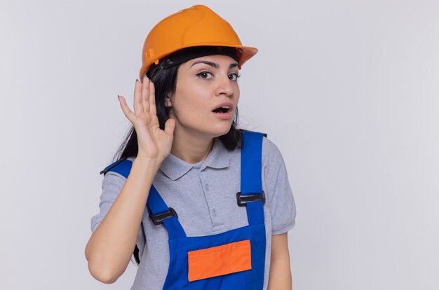 Young builder woman in construction uniform and safety helmet holding hand over her ear trying to listen to gossips standing over white wall