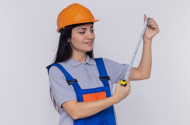 Young builder woman in construction uniform and safety helmet holding measure tape looking at it smiling standing over white wall