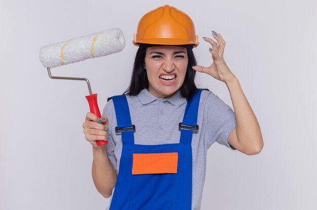 건설 유니폼 및 안전 헬멧에 젊은 작성기 여자 화가 전면을보고 페인트 롤러를 들고 흰색 벽 위에 서 제기 팔으로 화가