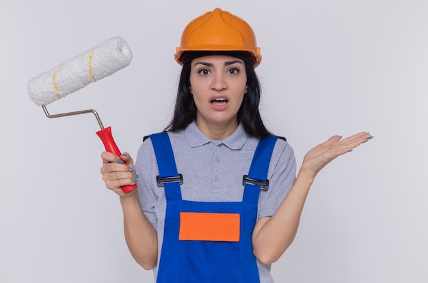 Young builder woman in construction uniform and safety helmet holding paint roller looking at front confused with arms raised standing over white wall