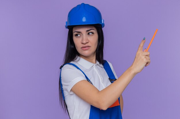 Young builder woman in construction uniform and safety helmet holding pencil pointing with it at something standing over purple wall