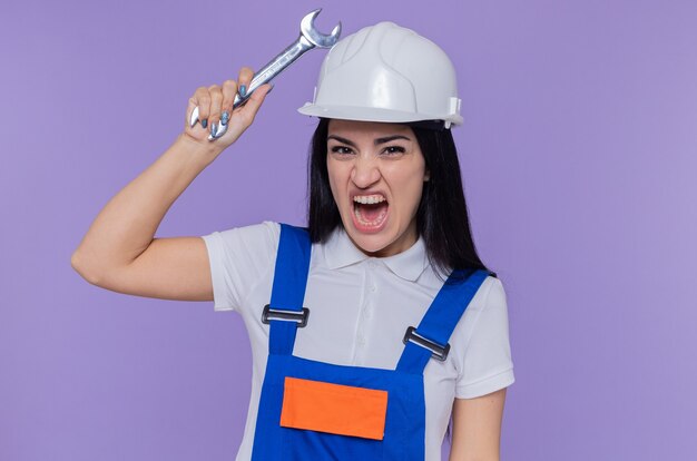 Young builder woman in construction uniform and safety helmet holding wrench looking at front shouting with aggressive expression standing over purple wall