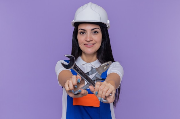 Young builder woman in construction uniform and safety helmet holding wrench and pliers looking at front crossing hands smiling confident standing over purple wall
