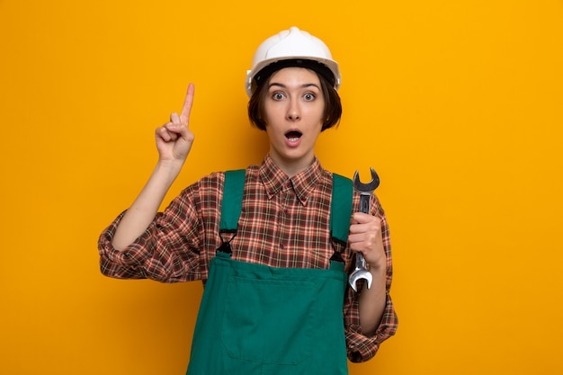 Foto gratuita giovane donna del costruttore in uniforme da costruzione e casco di sicurezza che tiene la chiave sorpresa che mostra il dito indice con una nuova idea in piedi sull'arancia