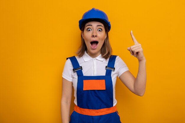 建設制服と安全ヘルメットの若いビルダーの女性は素晴らしいアイデアを持っている人差し指を示して驚いて驚いたように見えます