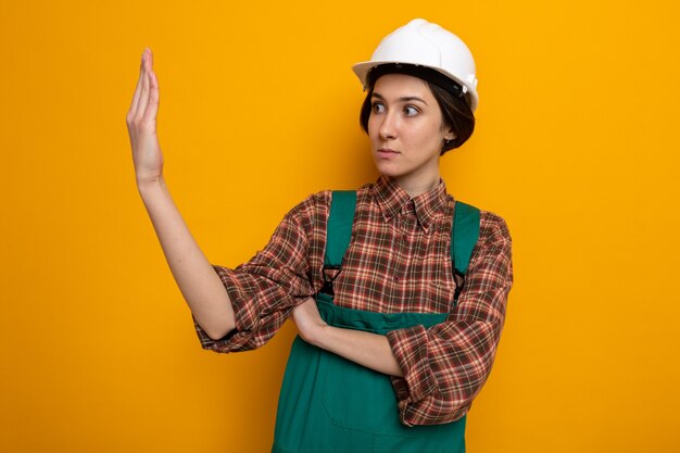 Молодая женщина-строитель в строительной форме и защитном шлеме, глядя в сторону на свою руку, обеспокоенная и смущенная, стоя на оранжевом