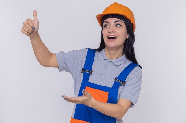 Молодая женщина-строитель в строительной форме и защитном шлеме, глядя в сторону, улыбаясь, показывая большой палец вверх, представляя с рукой, стоящей над белой стеной