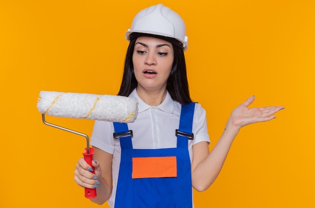 Young builder woman in construction uniform and safety helmet looking at confused