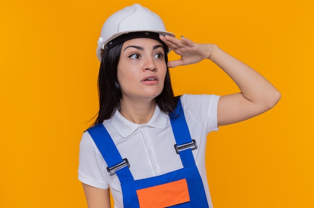 건설 유니폼 및 안전 헬멧에 젊은 작성기 여자 머리 위로 손으로 멀리 찾고 누군가 또는 오렌지 벽 위에 서있는 무언가를보고