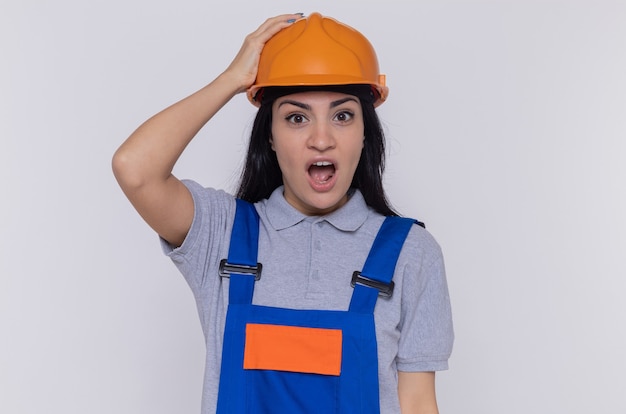 Young builder woman in construction uniform and safety helmet looking at front amazed and surprised standing over white wall