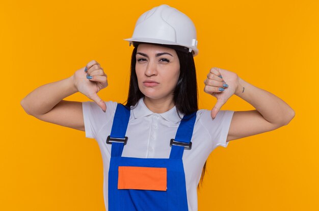 オレンジ色の壁の上に立って親指を下に見せて不快になっている正面を見て建設制服と安全ヘルメットの若いビルダーの女性