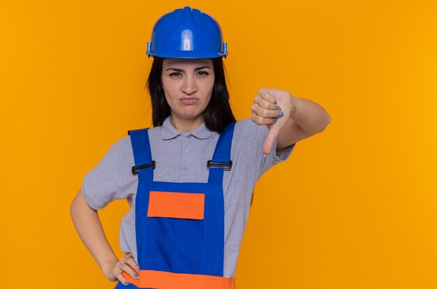オレンジ色の壁の上に立って親指を下に見せて不機嫌な正面を見て建設制服と安全ヘルメットの若いビルダーの女性
