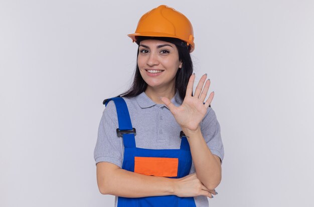 Young builder woman in construction uniform and safety helmet looking at front happy and positive showing open palm standing over white wall