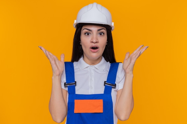 Молодая женщина-строитель в строительной форме и защитном шлеме, глядя на фронт, счастливые и удивленные, поднимая руки, стоя над оранжевой стеной