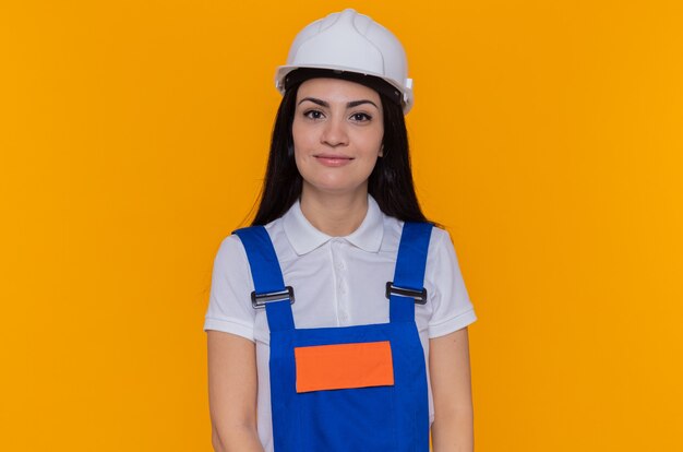 Free photo young builder woman in construction uniform and safety helmet looking at front smiling confident standing over orange wall