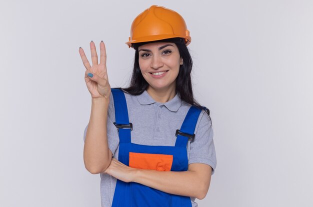 Молодая женщина-строитель в строительной форме и защитном шлеме, глядя на переднюю улыбку, показывающую номер три с пальцами, стоящими над белой стеной