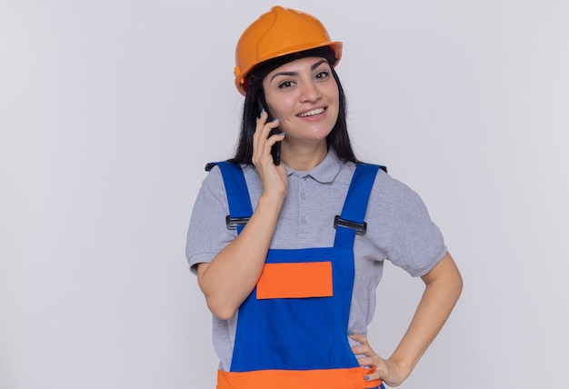 Giovane donna del costruttore in uniforme da costruzione e casco di sicurezza guardando davanti sorridente mentre parla al telefono cellulare in piedi sopra il muro bianco