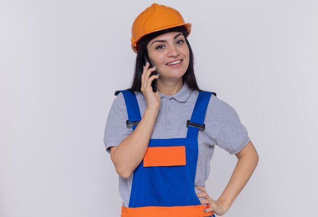 Young builder woman in construction uniform and safety helmet looking at front smiling while talking on mobile phone standing over white wall