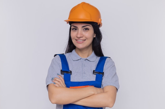 Young builder woman in construction uniform and safety helmet looking at front with confident expression smiling with arms crossed standing over white  wall