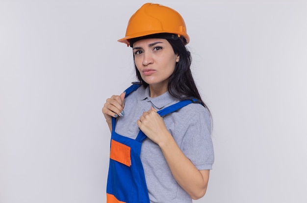 Молодая женщина-строитель в строительной форме и защитном шлеме смотрит вперед с серьезным уверенным выражением лица, стоящим над белой стеной