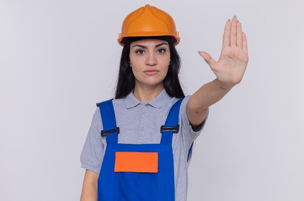 Молодая женщина-строитель в строительной форме и защитном шлеме, глядя вперед с серьезным лицом, делая жест остановки с рукой, стоящей над белой стеной