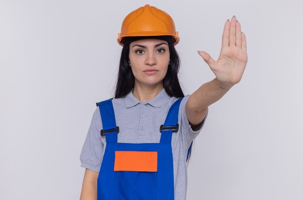 Young builder woman in construction uniform and safety helmet looking at front with serious face making stop gesture with hand standing over white wall