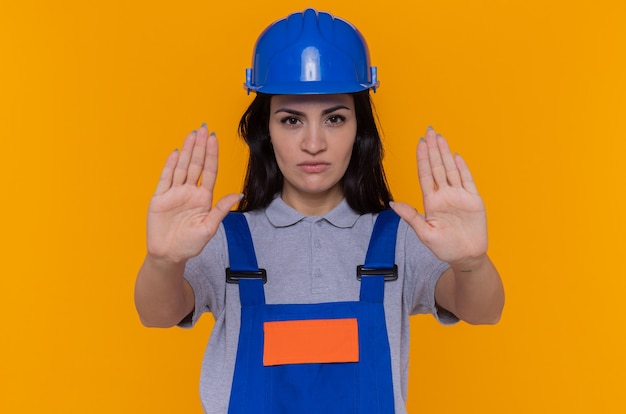 Free photo young builder woman in construction uniform and safety helmet looking at front with serious face making stop gesture with hands standing over orange wall