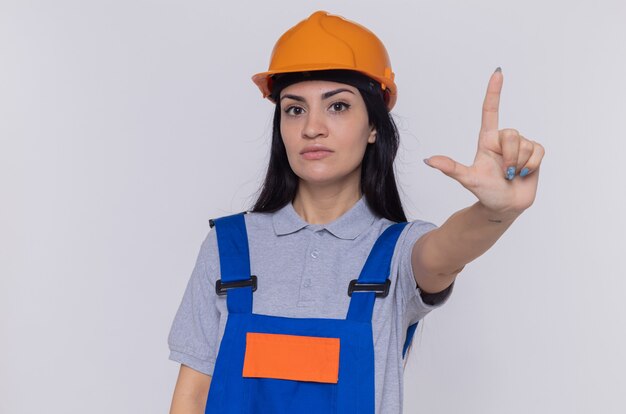 Young builder woman in construction uniform and safety helmet looking at front with serious face showing index finger warning standing over white wall