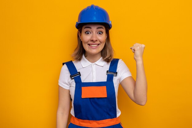 建設ユニフォームと安全ヘルメットの若いビルダーの女性は、勝者のように拳を上げて幸せで興奮しているように見えます