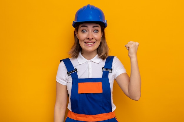 Молодая женщина-строитель в строительной форме и защитном шлеме выглядит счастливой и взволнованной, поднимая кулак, как победитель