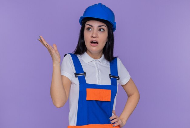 紫色の壁の上に立って腕を上げて混乱し、驚いて見上げる建設制服と安全ヘルメットの若いビルダーの女性