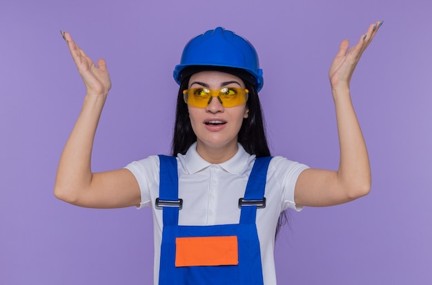 Young builder woman in construction uniform and safety helmet looking up happy and surprised with arms raised standing over purple wall