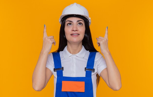 Young builder woman in construction uniform and safety helmet looking up smiling confident