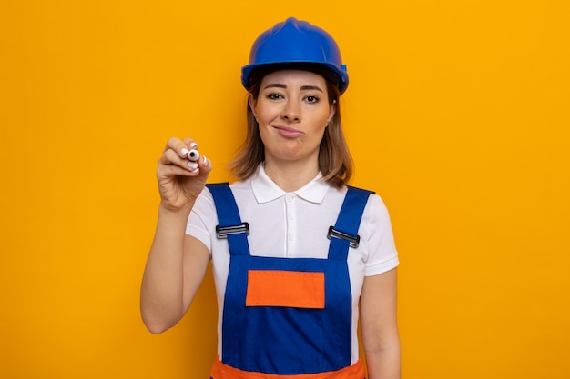 Молодая женщина-строитель в строительной форме и защитном шлеме смотрит со скептической улыбкой на лице и пишет ручкой в воздухе