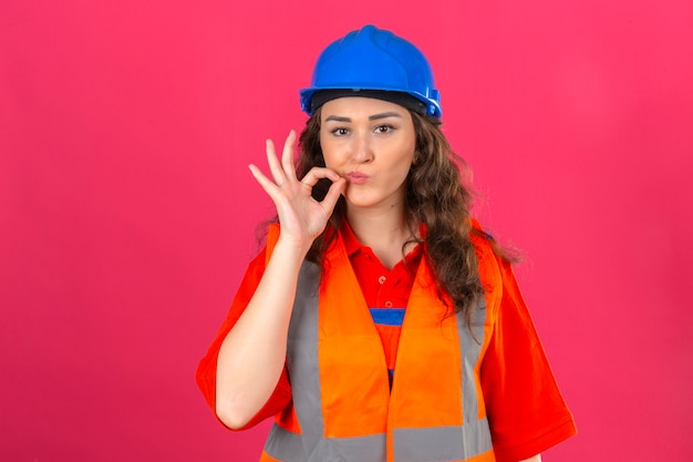 Young builder woman in construction uniform and safety helmet making silence gesture doing like closing her mouth with a zipper over isolated pink wall