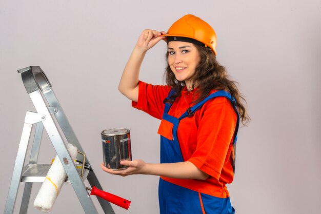 Young builder woman in construction uniform and safety helmet on a metal ladder with paint can smiling and touching her helmet over isolated white wall