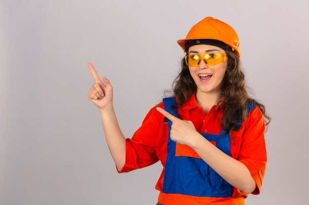 Молодой строитель женщина в строительной форме и защитный шлем, указывая пальцами в сторону с счастливым и смешным лицом на изолированной белой стене