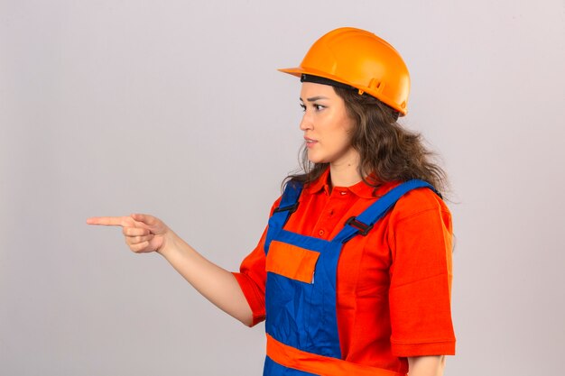 Giovane donna del costruttore in uniforme della costruzione e casco di sicurezza che indicano il lato con il dito indice che controlla parete bianca isolata