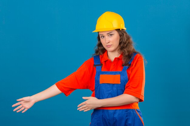 Young builder woman in construction uniform and safety helmet presenting and pointing with palms of hands looking at the camera with serious face over isolated blue wall