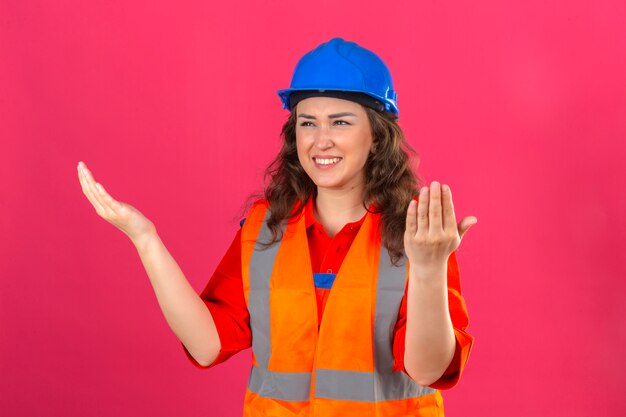 Young builder woman in construction uniform and safety helmet raising hands sideways in dismay and disappointment stare confused puzzled what happened over isolated pink wall