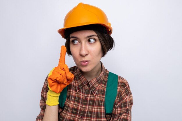 Young builder woman in construction uniform and safety helmet in rubber gloves looking aside with serious face showing index finger standing over white wall